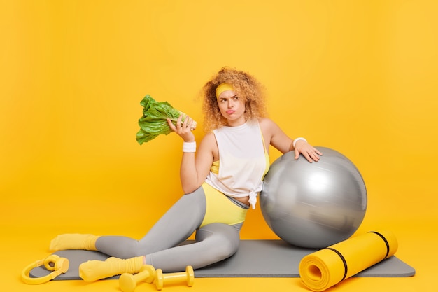 Woman dressed in activewear keeps to diet holds green vegetable poses on mat with sport equipment