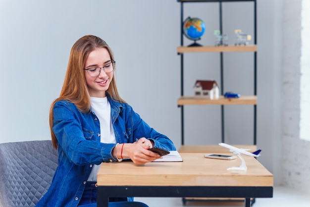 Woman dressed in denim shirt sits at the table during planning vacation trip uses smart phone.