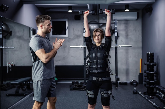 Woman dressed in an electric muscular suit lifts the dumbbells above her head