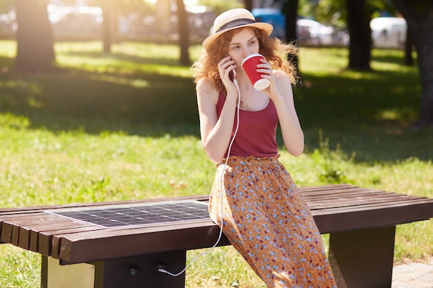 Woman dressed floor skirt, t shirt and hat, charging smart phone via USB outdoors. Public charging on bench with solar panel on street. Alternative electricity source and modern technology concept.