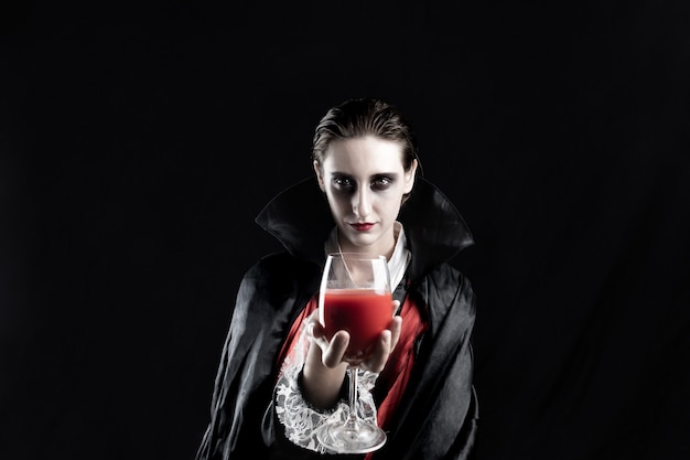 赤い飲み物のガラスを保持しているハロウィーンの吸血鬼として服を着た女性。黒の背景にドラキュラ衣装の若い女性の劇的なライトで撮影スタジオ