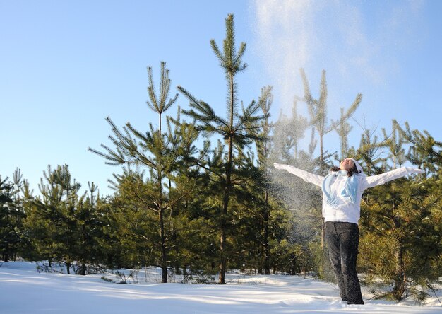 Woman dressed in winter clothes, stands among pine trees and throws up snow. Full height photo, hands raised up