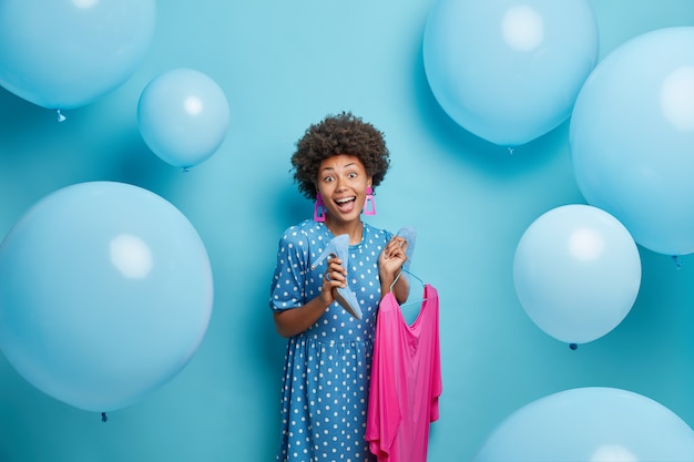 woman dresses for party holds pink dress on hangers and fasionable high heeled shoes poses on blue