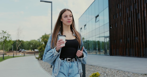 Женщина пьет кофе во время прогулки по улице с планшетом Современный образ жизни людей в городе Молодая женщина гуляет по центральному деловому району современного города