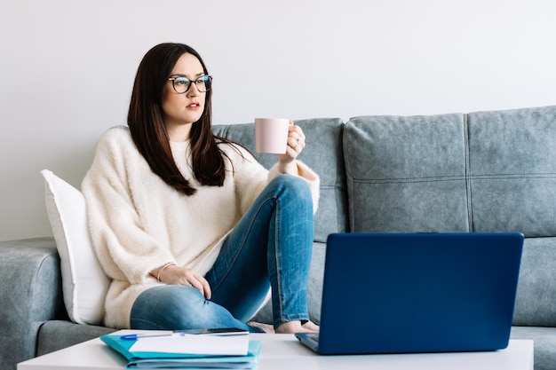 ノートパソコンで作業しながら自宅でコーヒーを飲む女性