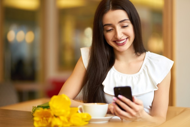 コーヒーを飲みながら電話を使う女性。春のスタイル。素敵な春のブルネットの女性。カフェで春のチューリップの花の花束と美しい若い女性。美しい春の気分