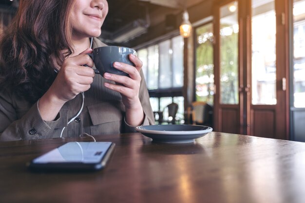 カフェで携帯電話とイヤホンで音楽を聴きながらコーヒーを飲む女性