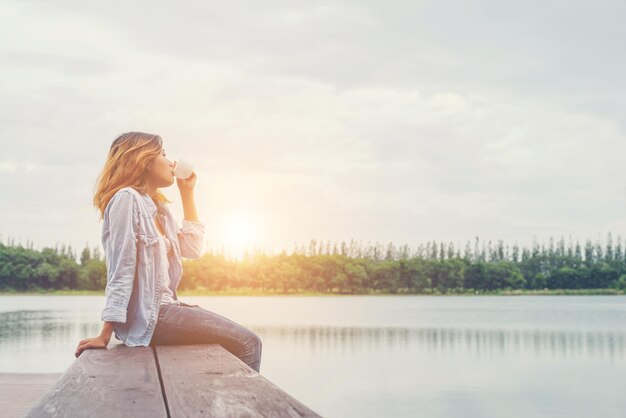 写真 夕暮れ中に湖の上の埠頭に座ってコーヒーを飲んでいる女性