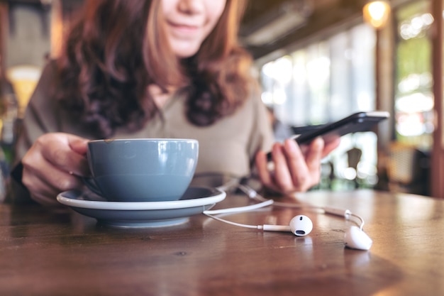 カフェの木製テーブルでイヤホンで音楽を聴きながら携帯電話を使用しながらコーヒーを飲む女性