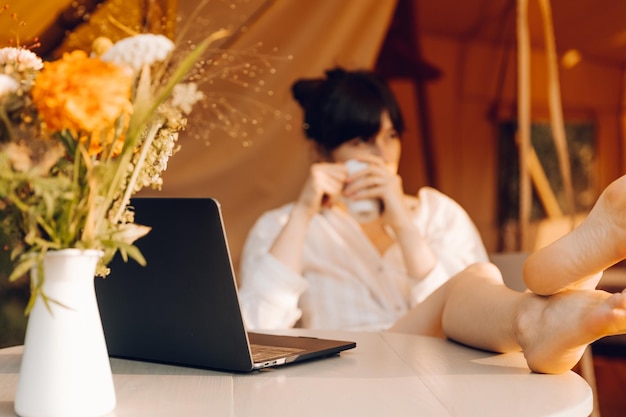 a woman drinking from a cup next to a laptop and a vase of flowers