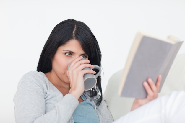 Woman drinking from a grey mug while reading a book