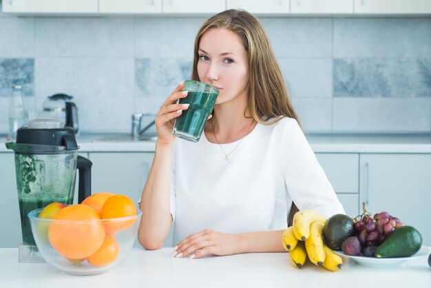 白いキッチンで緑のスピルリナを飲む女性は、完璧なボディとカロリービーガンの食事をデトックスします...