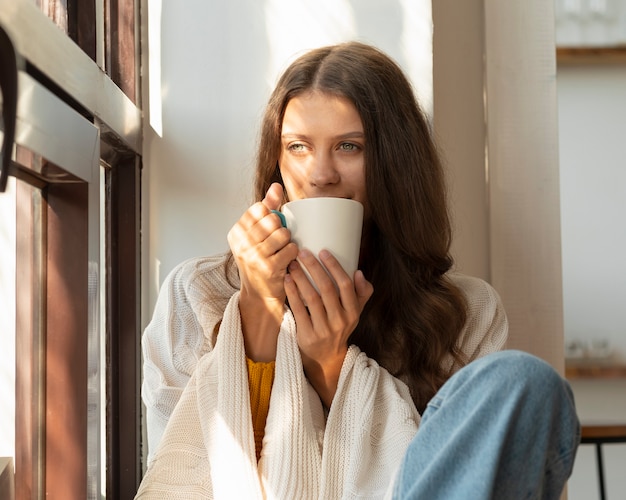 熱い飲み物を飲み、朝を楽しんで、窓に座って夢を見ている女性