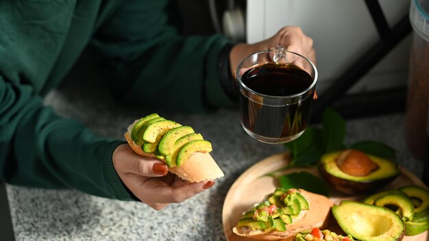 Woman drinking hot coffee and eating avocado sandwiches in kitchen