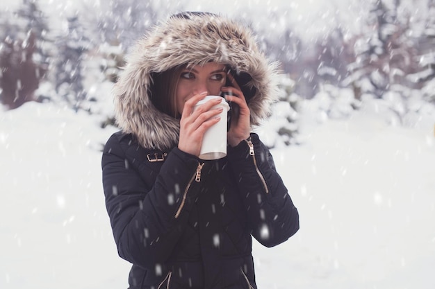 写真 冬の雪片の下でマグカップからホットコーヒーやお茶を飲む女性