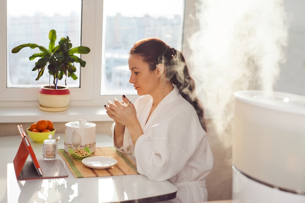 Woman drinking tea reading tablet near humidifier