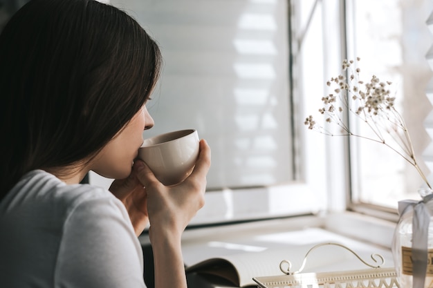 晴れた朝に窓際に座って、お茶を飲む女性。家で休む。