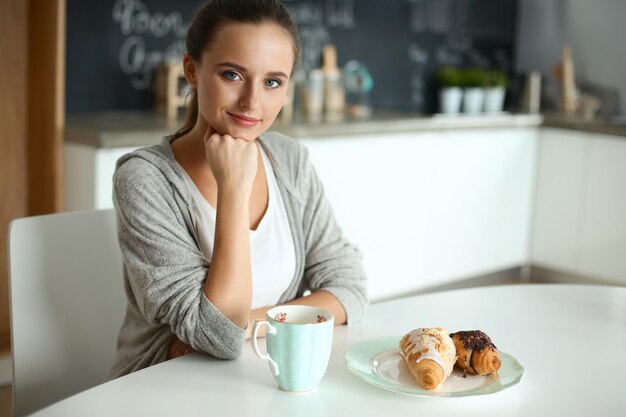 台所のテーブルで甘いクロワッサンとお茶を飲む女性