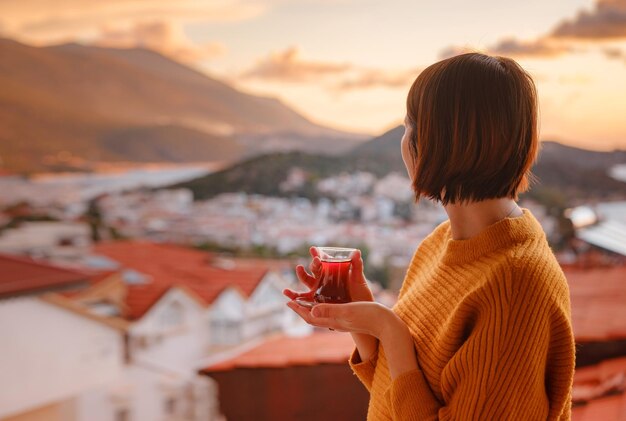 Woman drinking turkish tea from traditional turkish teacup and enjoys panorama over sunset of Kas resort town of Mediterranean sea in Turkey