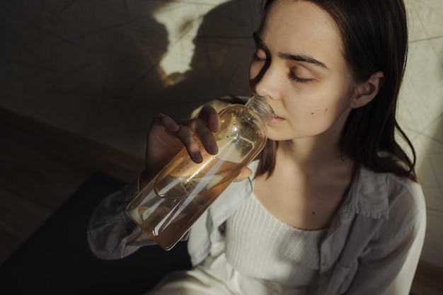 woman drinking water from glass bottle during yoga session at home in room lit by sunlight