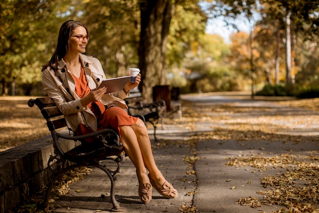 Женщина пьет кофе и сидит на скамейке в парке в осеннюю погоду, используя планшетный пк и проверяя социальные сети.