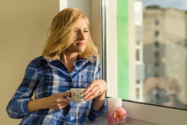 Woman drinks coffee, looks out the window at home