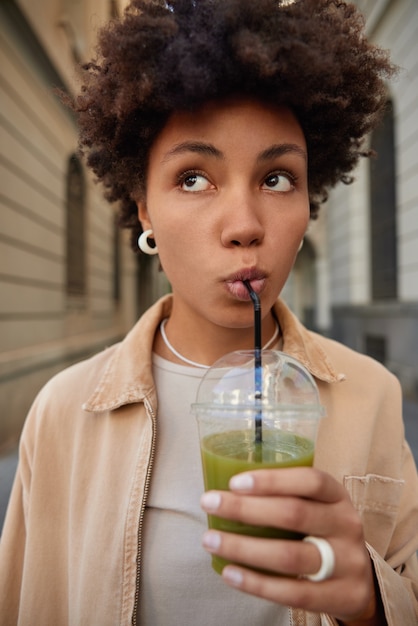 写真 女性はストローからプラスチックカップで天然の緑のスムージーを飲みますスタイリッシュな服を着て街の外を歩き、ビーガンローフードを続けます