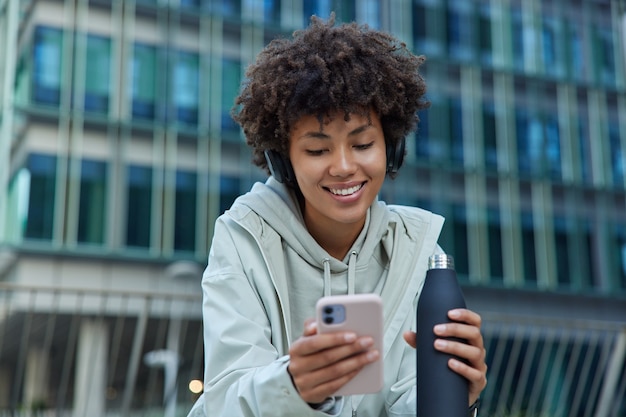 woman drinks water after exercising uses mobile phone scrolls social networks smiles happily dressed in hoodie and anorak poses against modern city building enjoys messaging