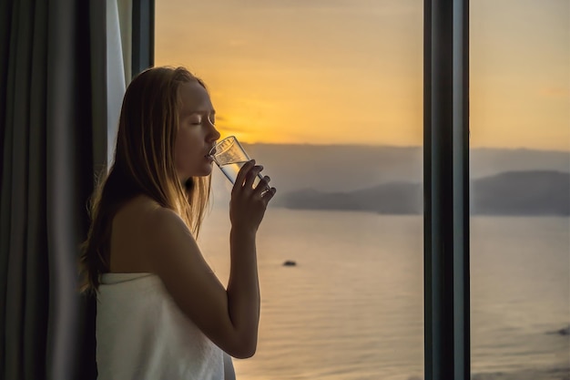 Женщина пьет воду утром на фоне окна с видом на море