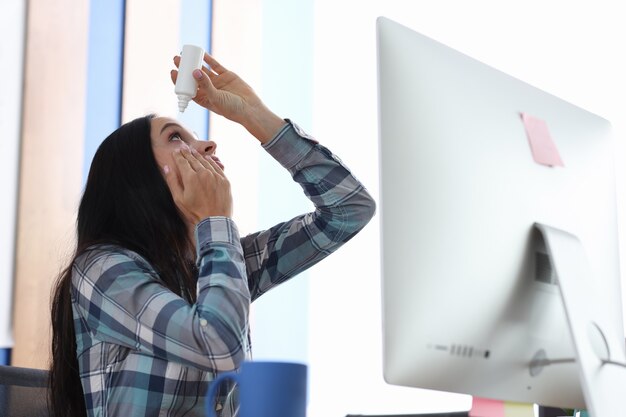 Woman drips eye drops into eyes while sitting at workplace at computer