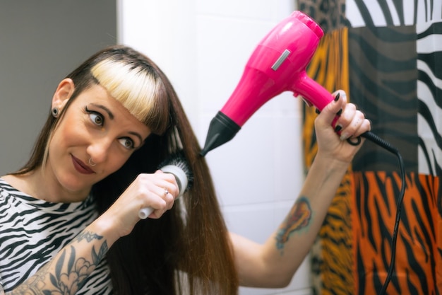 写真 バスルームの鏡の前で染めた髪を乾かす女性。カジュアルルックのピアスとタトゥー