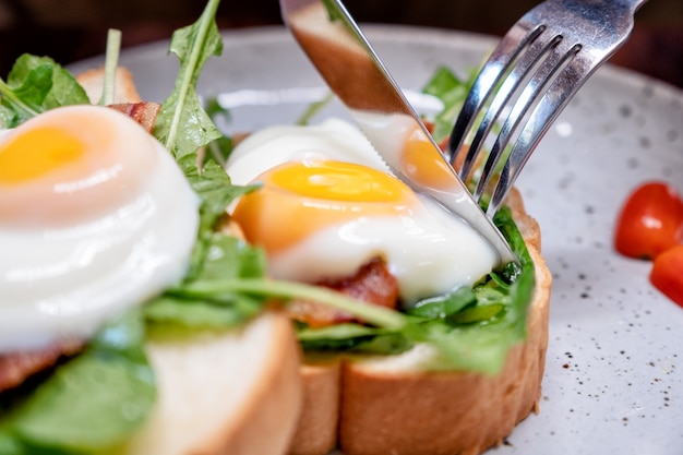 A woman eating breakfast sandwich with eggs, bacon and sour cream by knife and fork in a plate on wooden table