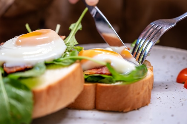 A woman eating breakfast sandwich with eggs, bacon and sour cream by knife and spoon in a plate on wooden table