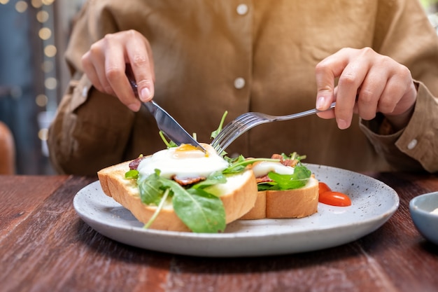 木製のテーブルの上の皿にナイフとスプーンで卵、ベーコン、サワークリームと朝食サンドイッチを食べる女性