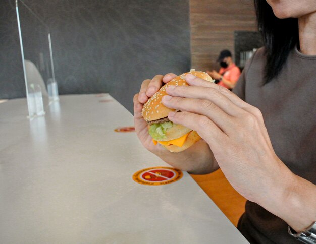 코로나바이러스 코비드-19, 레스토랑 및 사회적 거리 개념으로부터 감염을 보호하기 위해 테이블 실드 플라스틱 칸막이가 있는 음식을 먹는 레스토랑에서 햄버거를 먹는 여성