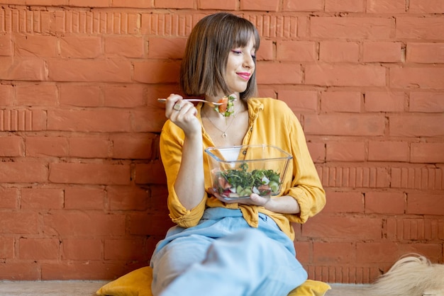 Woman eating healthy salad while sitting on brick wall background