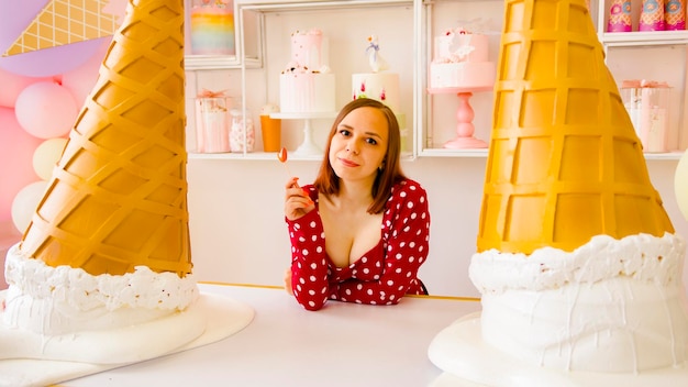 Woman eating lollipop in confectionery Content female looking at camera while eating sweet lollipop on table with large decorative ice cream cones in light confectionery