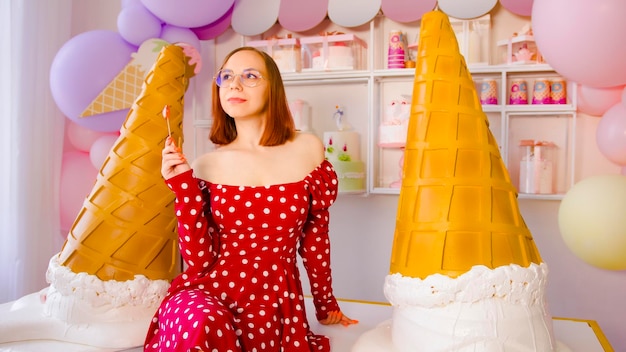 Woman eating lollipop in confectionery Content female looks away while eating sweet lollipop on table with large decorative ice cream cones in light confectionery