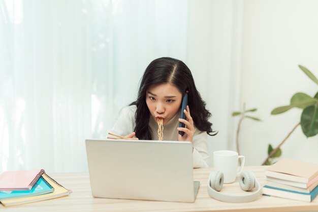 Photo woman eating noodles while working with laptop and cellphone in office