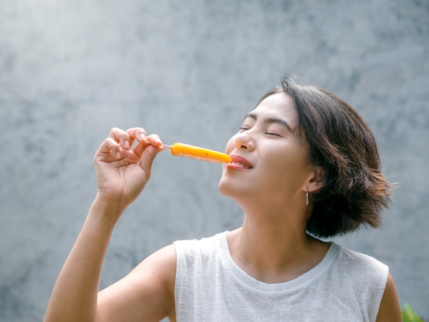 Woman eating popsicles. Happy beautiful Asian woman wearing casual white sleeveless shirt holding yellow popsicle, outdoors. Smiling female enjoying ice lolly in summer.