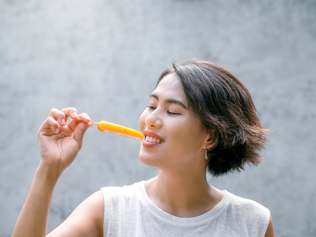 アイスキャンデーを食べる女性。屋外で、黄色のアイスキャンデーを保持しているカジュアルな白いノースリーブシャツを着て幸せな美しいアジアの女性。夏にアイスキャンデーを楽しんでいる笑顔の女性。