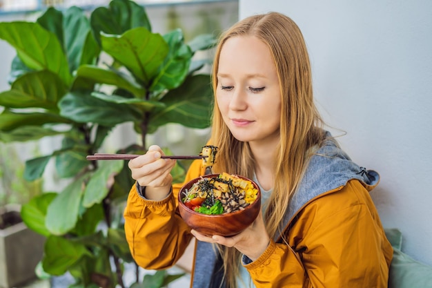 Woman eating raw organic poke bowl with rice and veggies closeup on the table top view from above