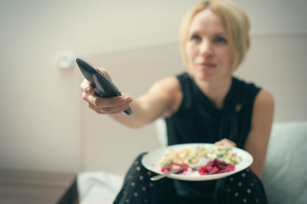 A woman eats in bed and holds a TV remote control