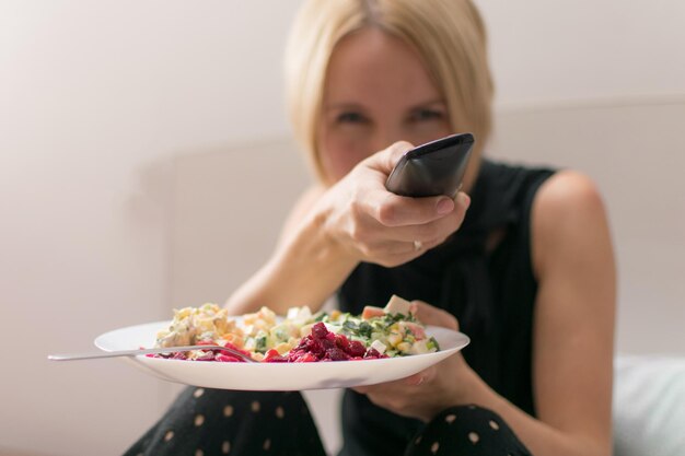 Photo a woman eats in bed and holds a tv remote control