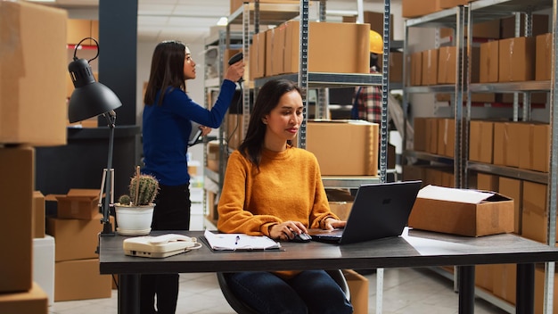 Woman employee analyzing products from racks and shelves, looking at supplies in cardboard packs before shipping order. Young owner working in storage room with business plan.