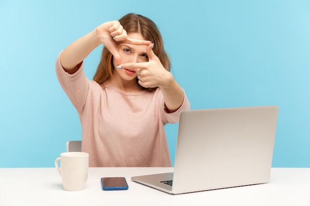 Photo woman employee designer or photographer sitting at workplace with laptop making photo frame with hands focusing at camera and cropping capturing business idea indoor studio shot blue background