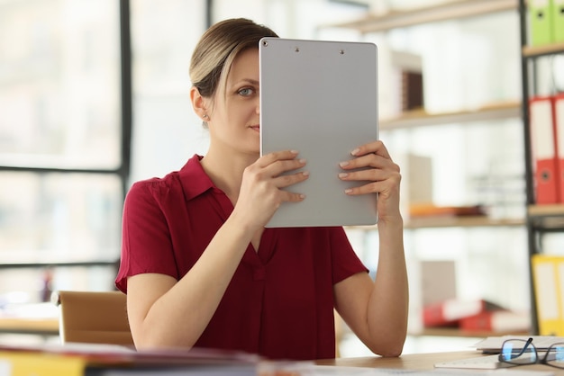 Woman employee lifts clipboard holding in hands hiding face