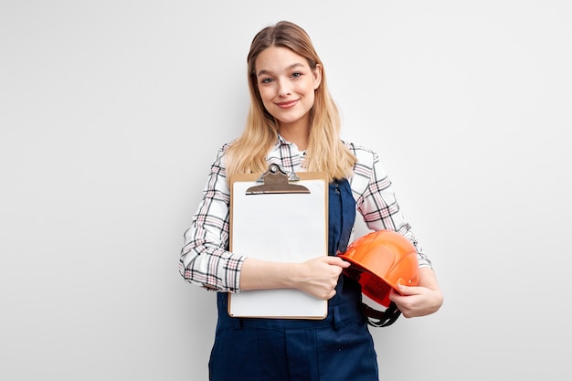 Женщина-инженер, держащая бумажную таблетку и шлем, одетая в униформу комбинезона построителя и смотрящую на камеру изолированную над белой предпосылкой студии.