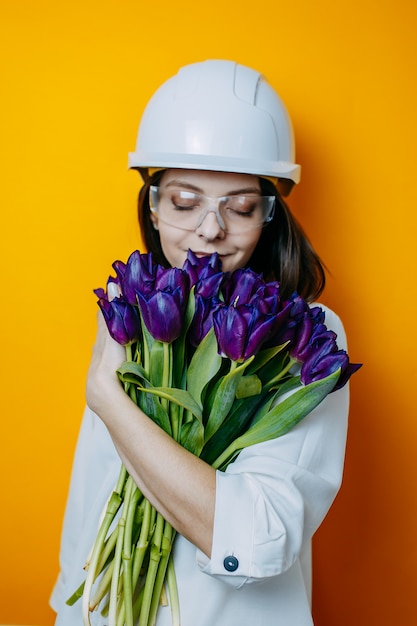 Woman engineer in white helmet and glasses. Safety poster. Woman in white shirt holds big bunch of purple tulips.