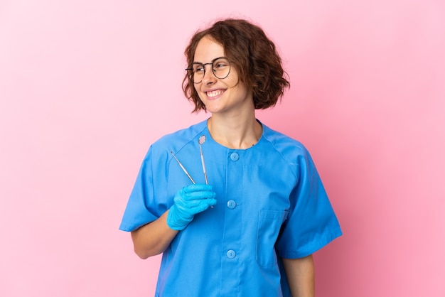 Woman English dentist holding tools over isolated on pink wall looking to the side and smiling